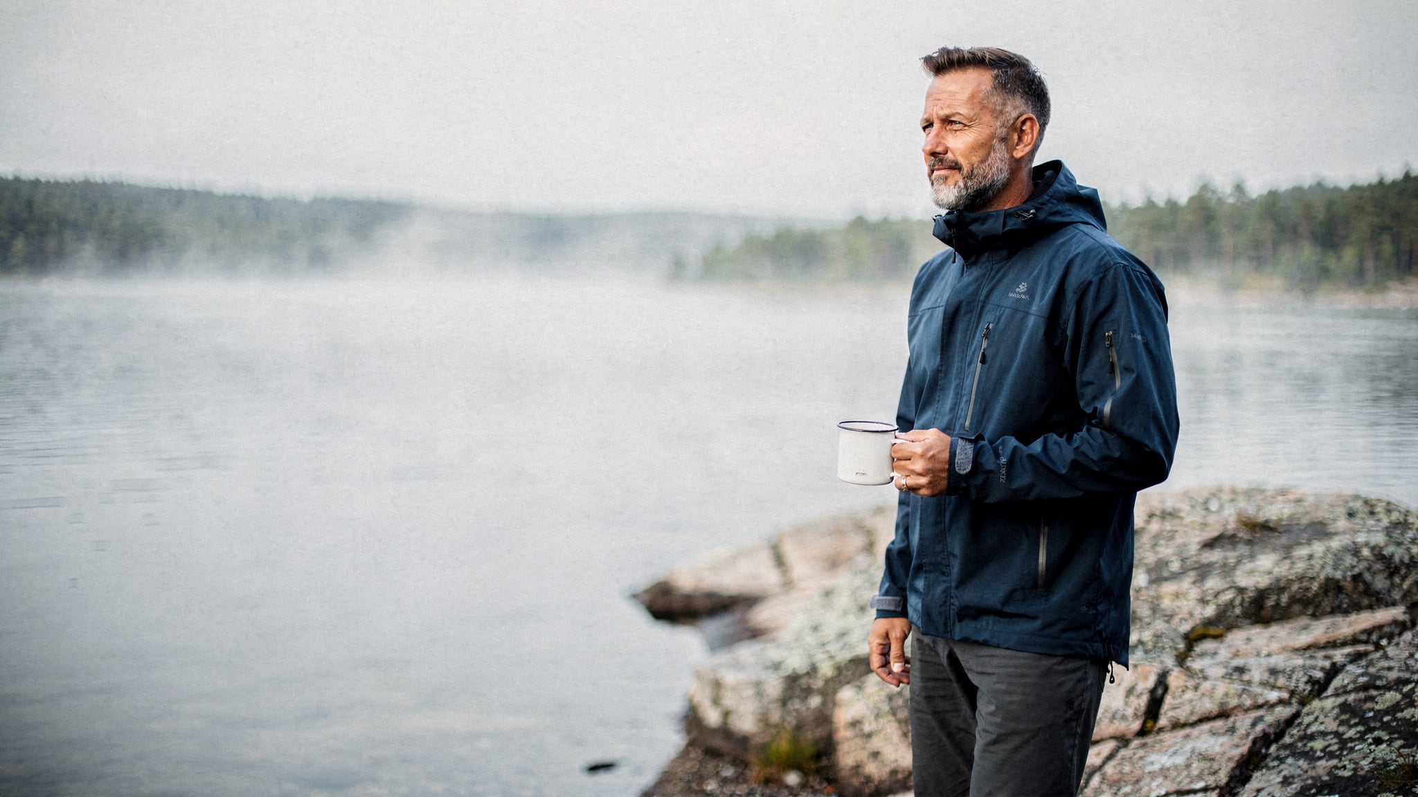 A man in his late forties in a blue outdoor jacket stands calmly on a granite rock beside a misty lake at dawn, holding a warm drink